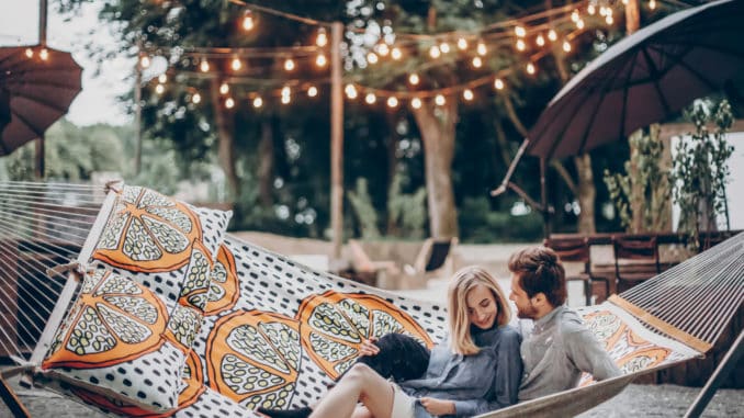 Stylish hipster couple cuddling and relaxing in hammock under retro lights in evening summer park.