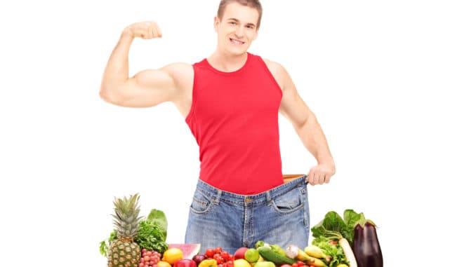 Weight loss man showing his muscles and standing behind a pile of fruits and vegetables, isolated on white background