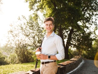 Happy young man in shirt walking at the city park with cup of coffee