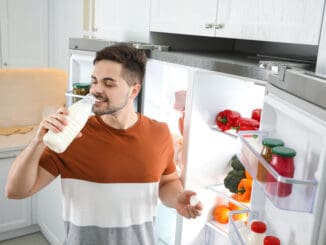 Young man drinking milk near open refrigerator in kitchen