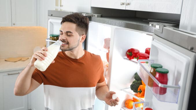 Young man drinking milk near open refrigerator in kitchen