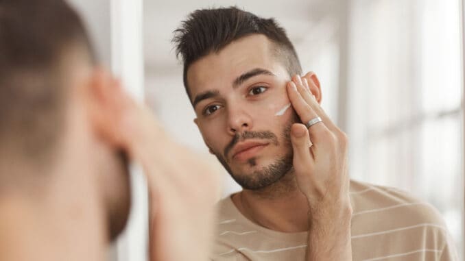 Portrait of young man applying face cream while looking in mirror during morning skincare routine