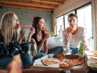 Three friends are sitting on sofa and eating pizza in house